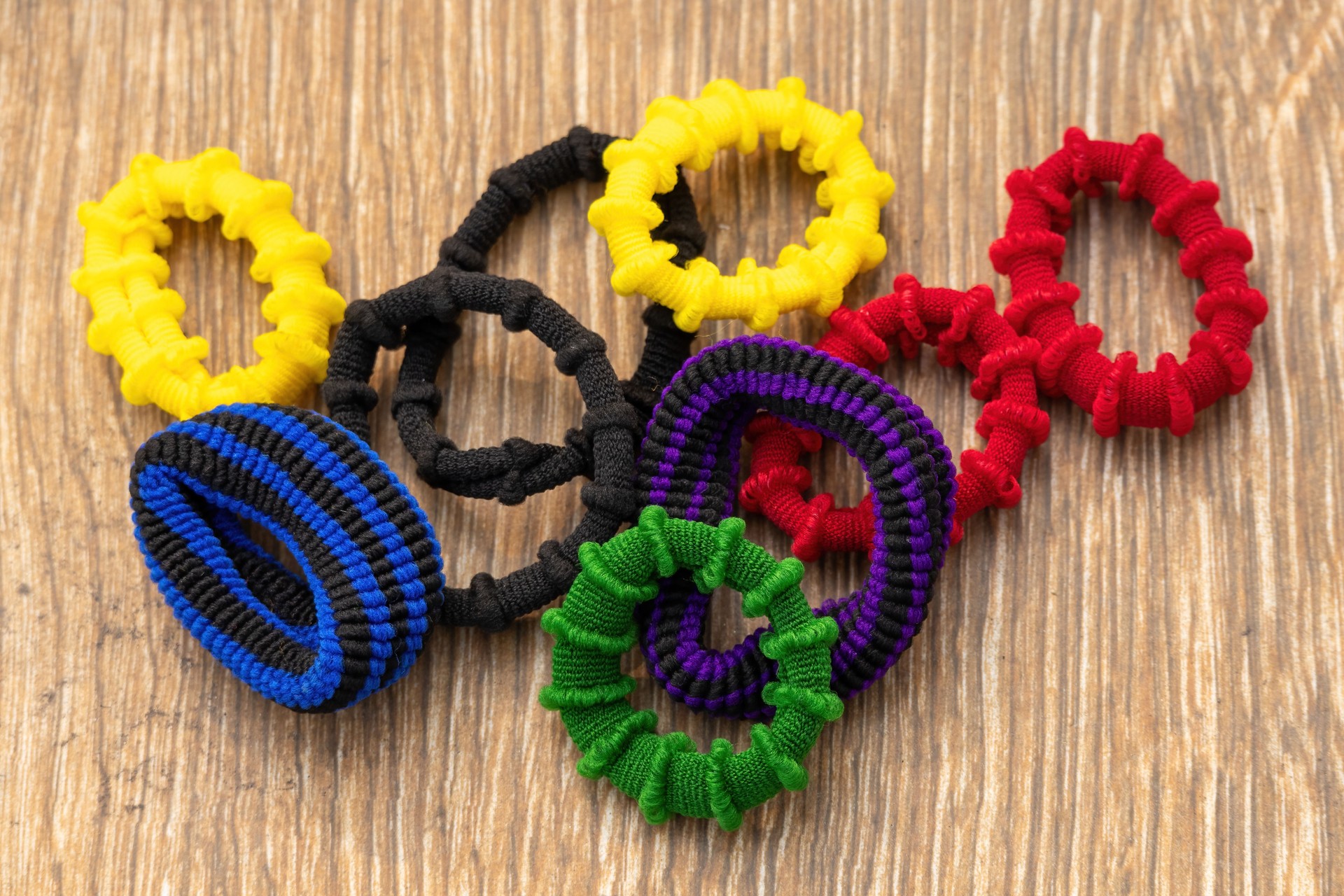 A collection of colorful scrunchies displayed on a wooden background. These are fabric-covered elastic hair ties used to fasten women's hair.
