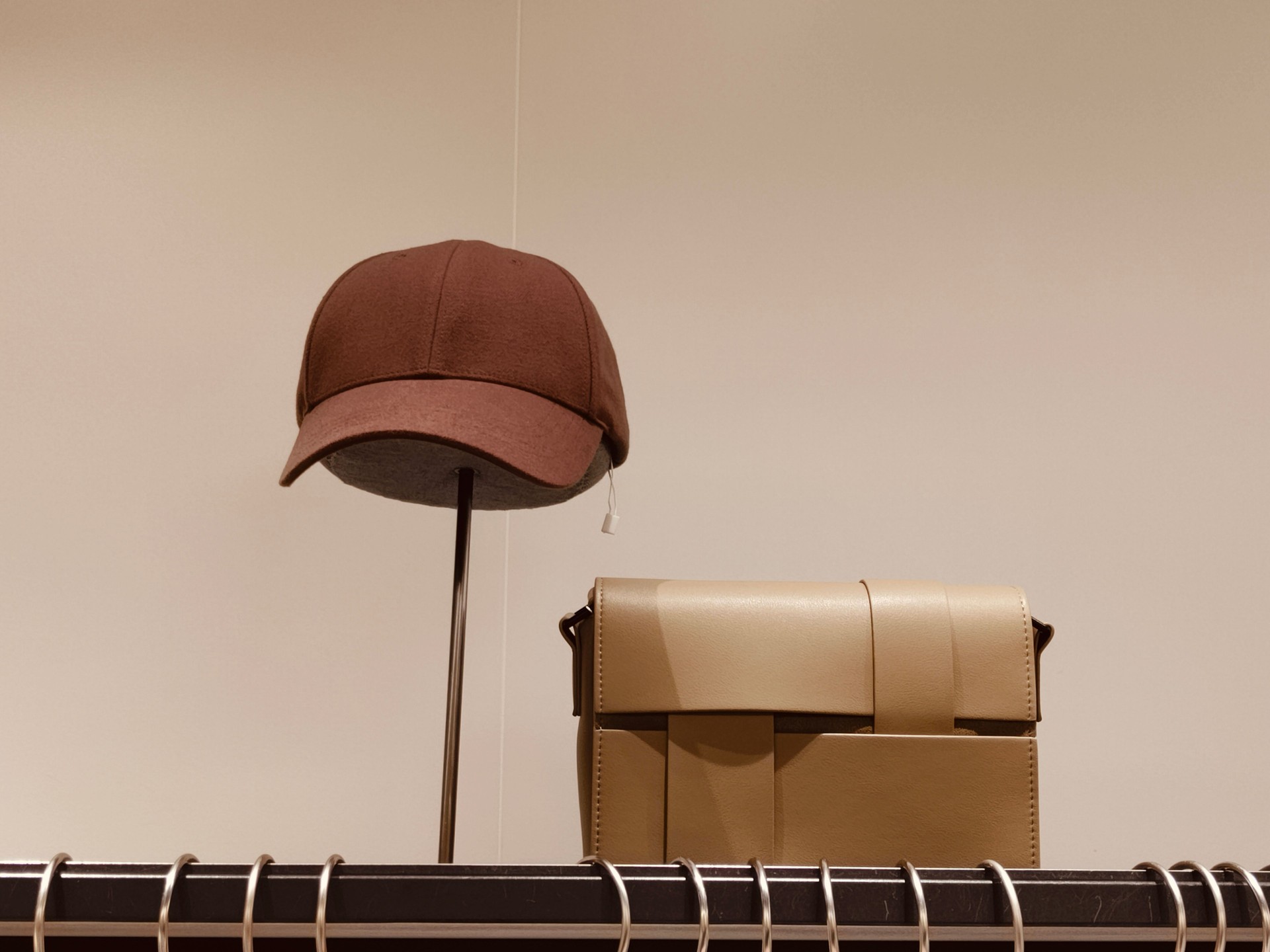 A brown baseball cap and a beige bag on top of the rack, in front of a white wall, a minimalist store display