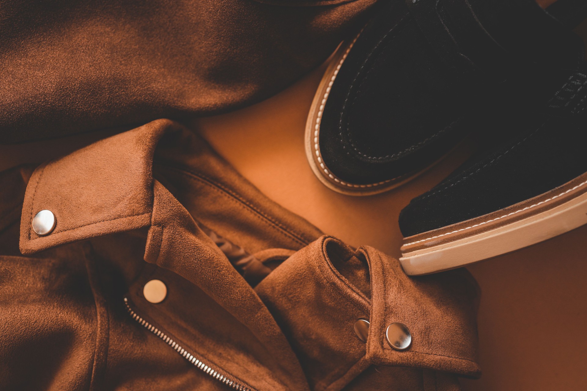 Flat lay of stylish brown suede jacket with metallic buttons and black suede shoes on a warm brown background, showcasing an elegant and modern autumn fashion outfit