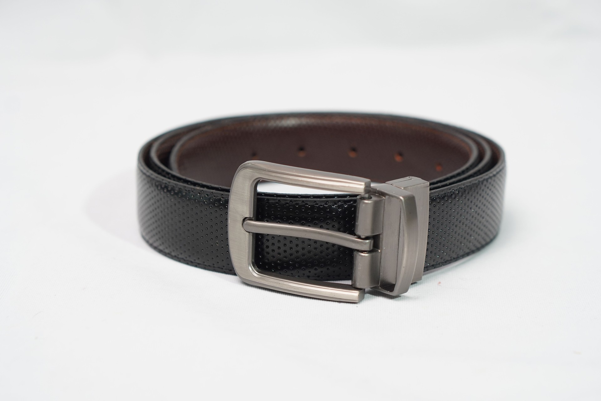 Coiled black perforated leather belt with a sleek gunmetal rotating buckle, offering versatile formal or casual style.