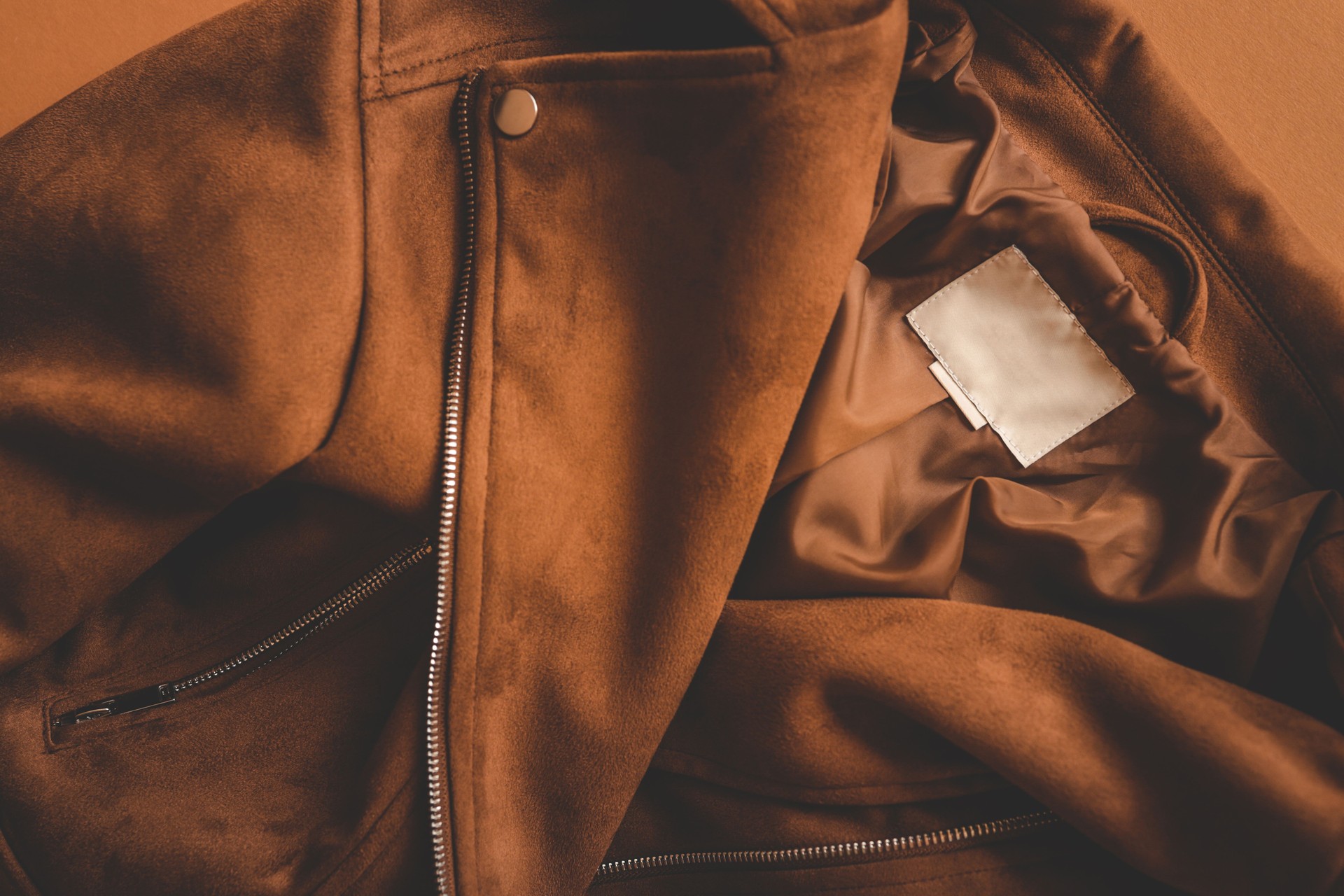 Close-up of a brown suede jacket with zipper details, showing the silky inner lining and a blank white label, highlighting the texture and craftsmanship of the garment